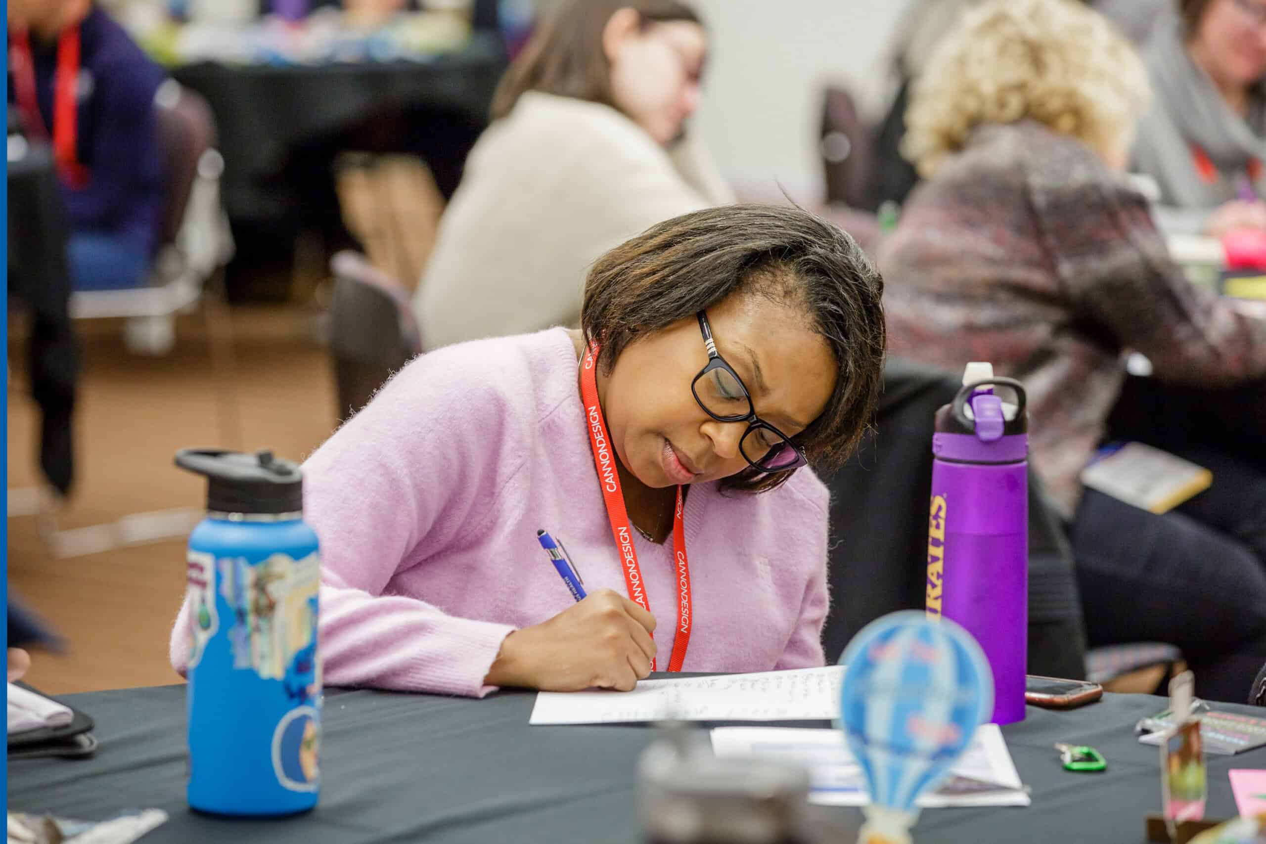 Person sitting at a table during a workshop, writing in a notebook with a pen, with others working in the background.