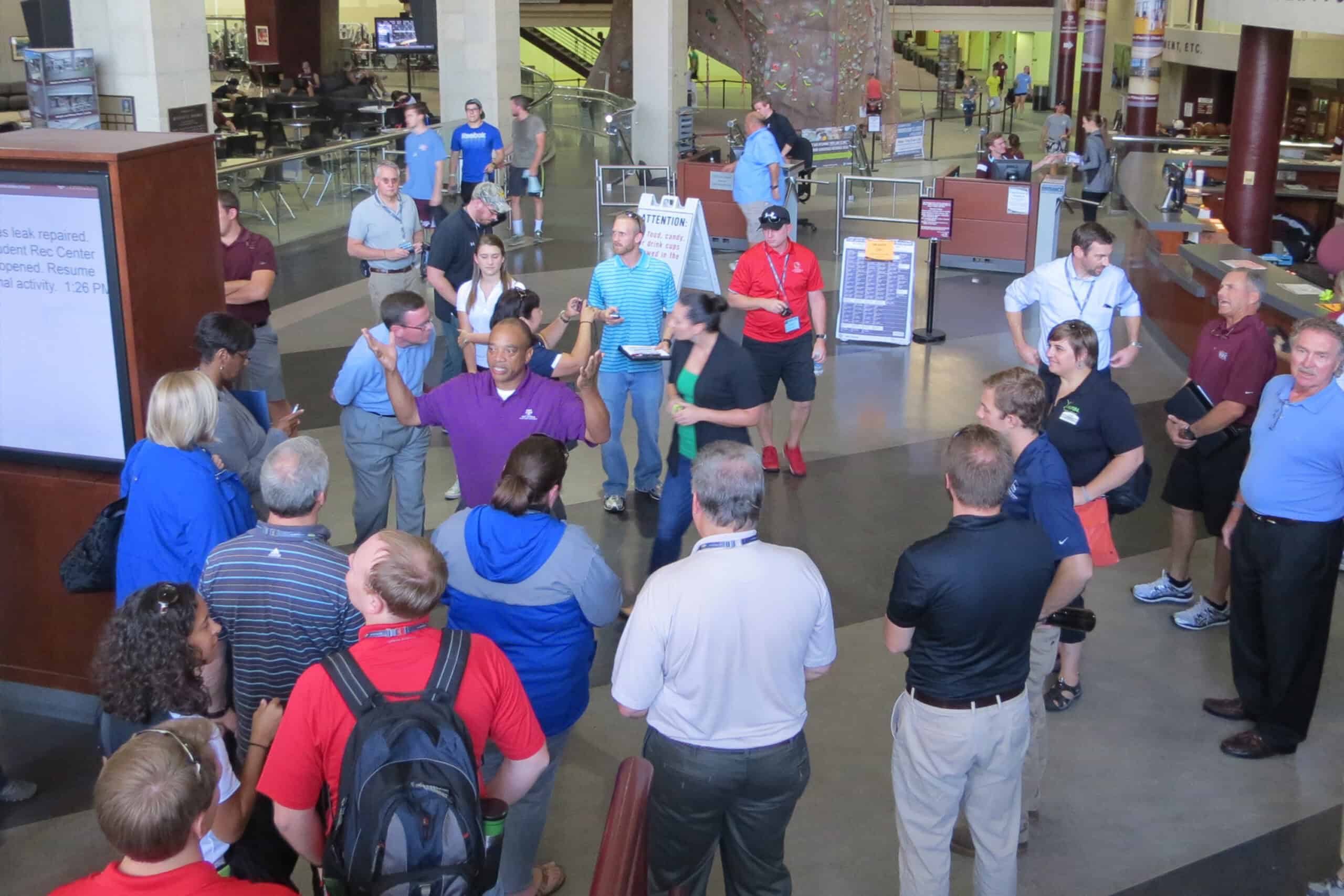 A group of people stands in a recreation center lobby while a guide speaks and gestures during a facility tour.
