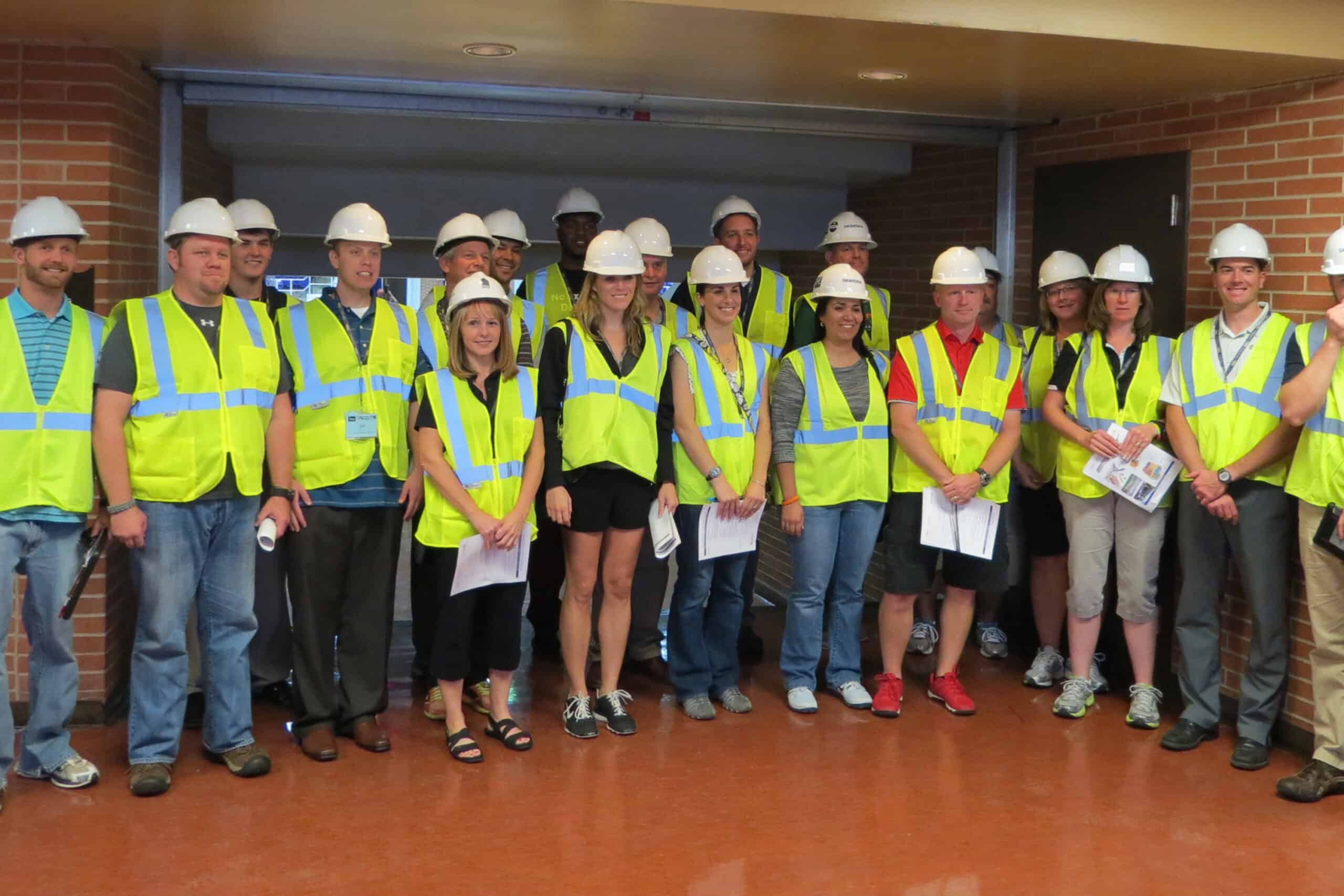 A group of people wearing hard hats and bright safety vests stands together indoors for a construction tour photo.