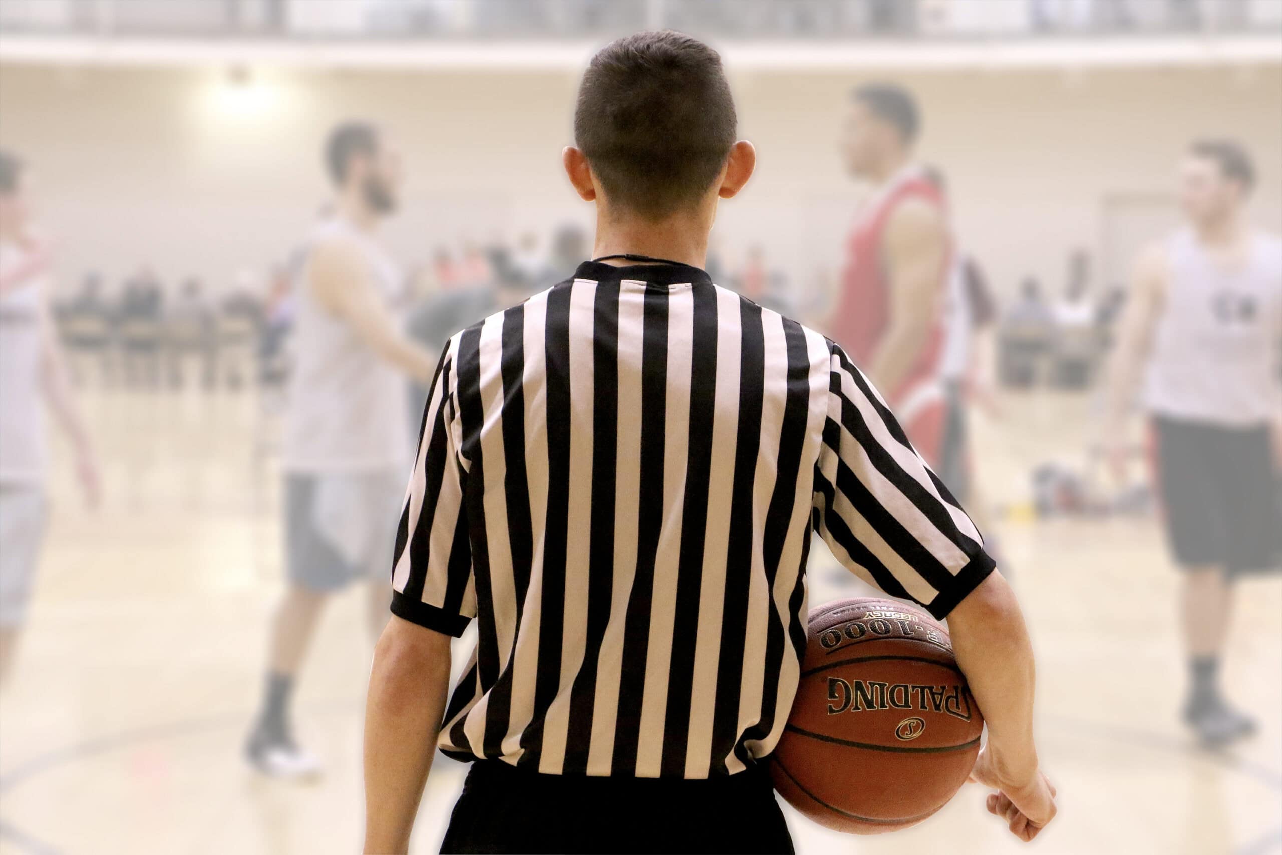 A referee photographed from behind with a basketball under his right arm.