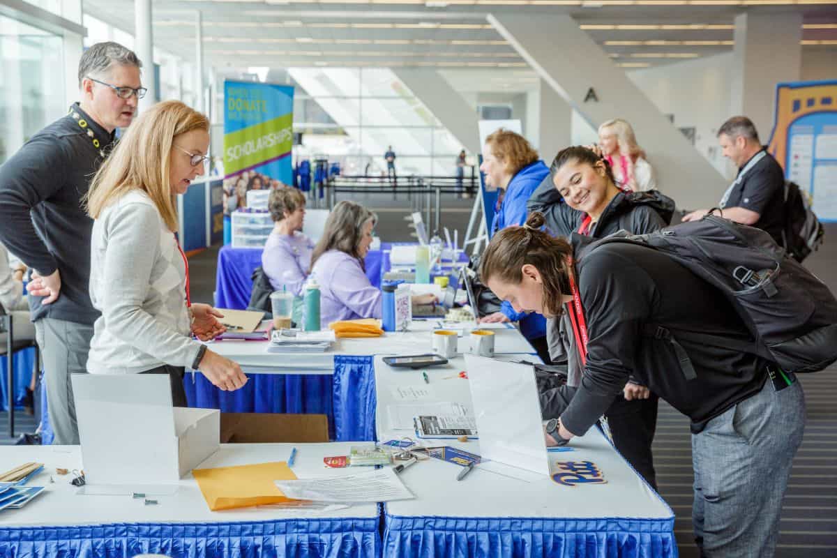 People at a Conference information table inside a large convention center, speaking with staff and filling out forms.