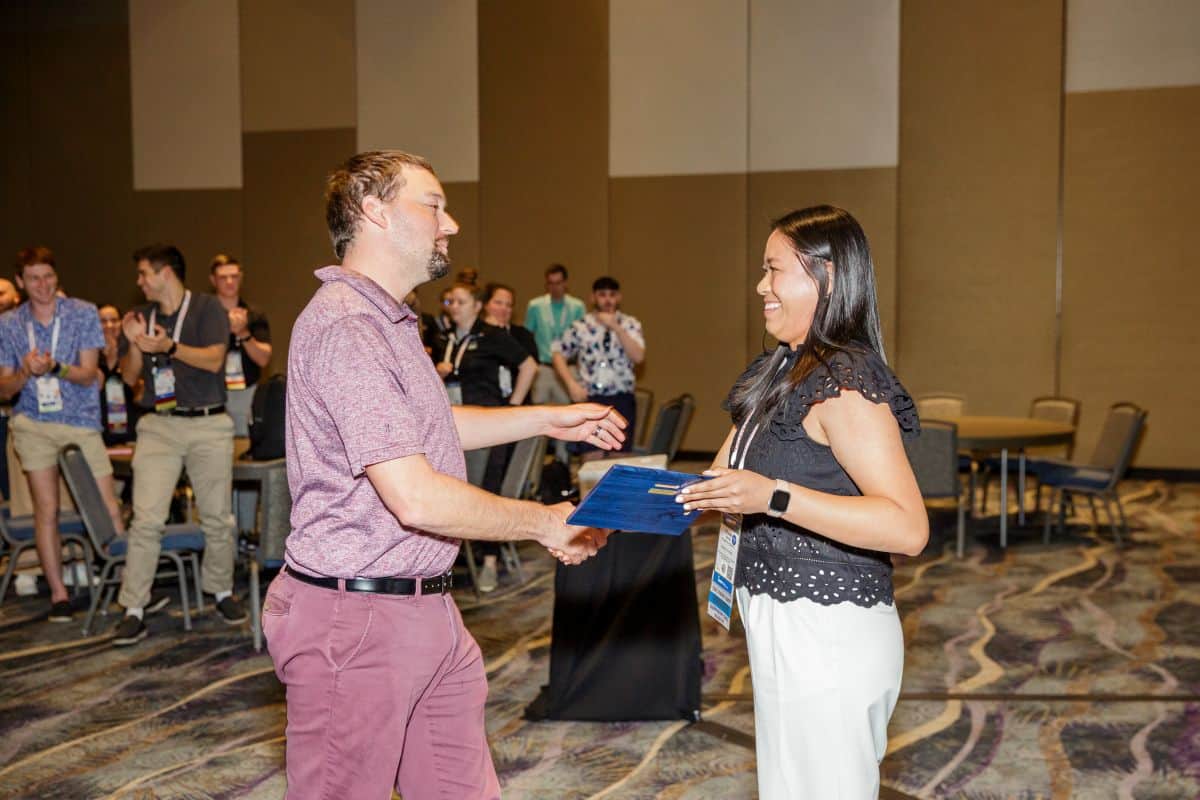 Two people facing each other in room shaking hands as a blue plaque is handed off. Several people in the background are standing and applauding.