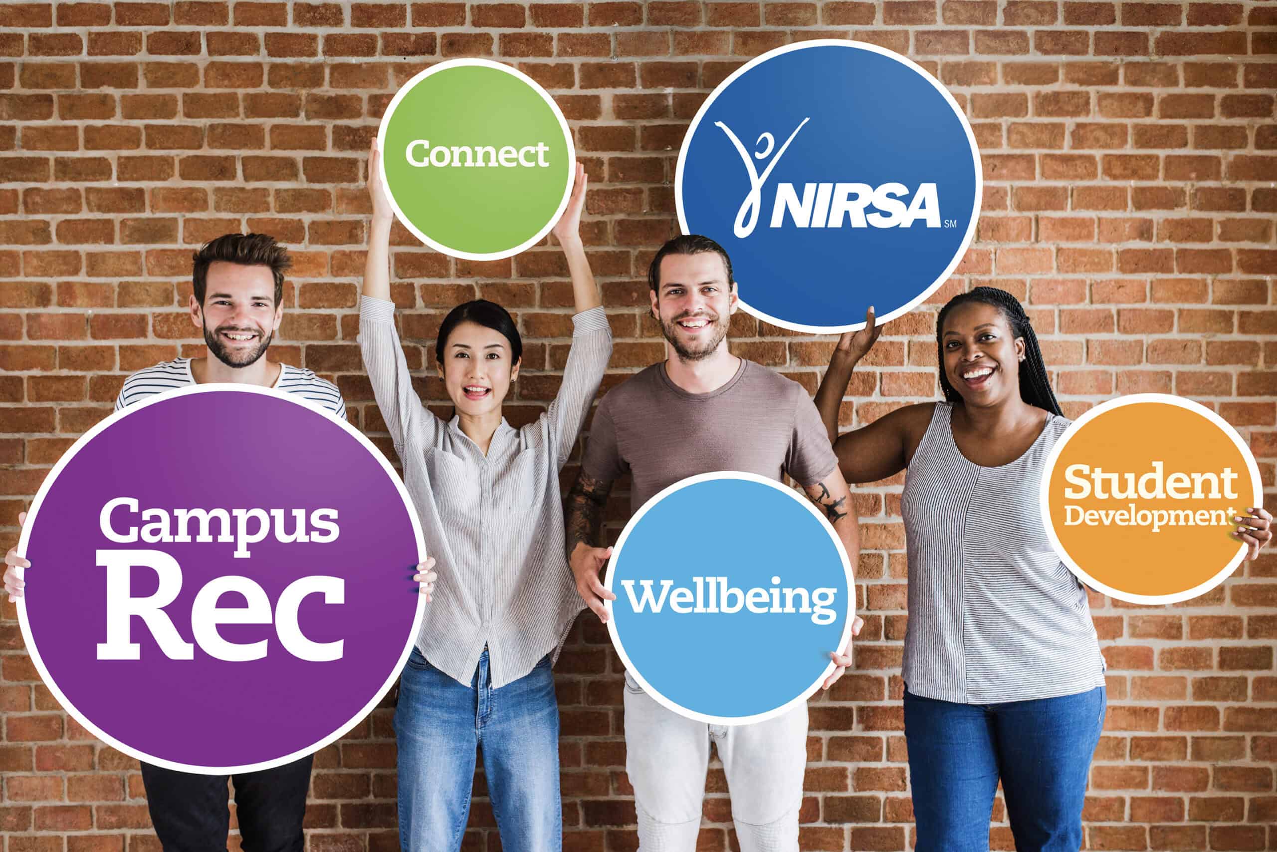 Diverse group of students holding signs for NIRSA, campus recreation, wellbeing, student development, and connection against brick wall.